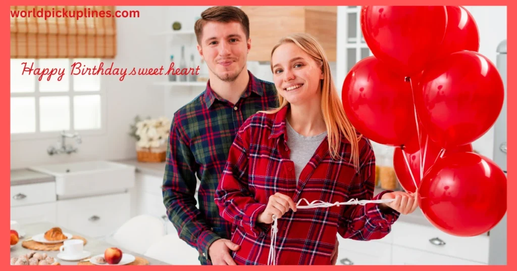 A young couple smiling together in a kitchen while the woman holds a bunch of red balloons.