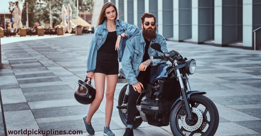 A bearded man sitting on a motorcycle beside a woman holding a helmet, both wearing matching denim jackets outdoors.