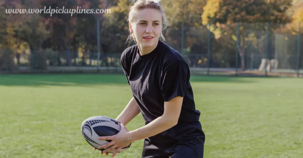 A female rugby player in a black shirt holds a rugby ball on a sunny grass field.