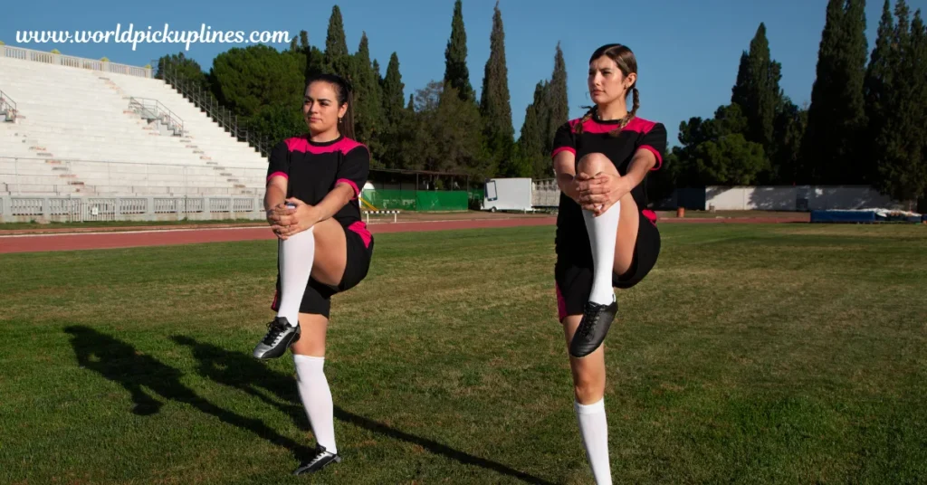 Two female soccer players in black and pink jerseys warm up by lifting one knee to their chest on a grassy field.